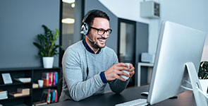 Mann mit Brille sitzt lachend im Büro am Schreibtisch vor einem Computermonitor. Er hält eine weisse Tasse in der Hand und hat Kopfhörer auf dem Kopf.